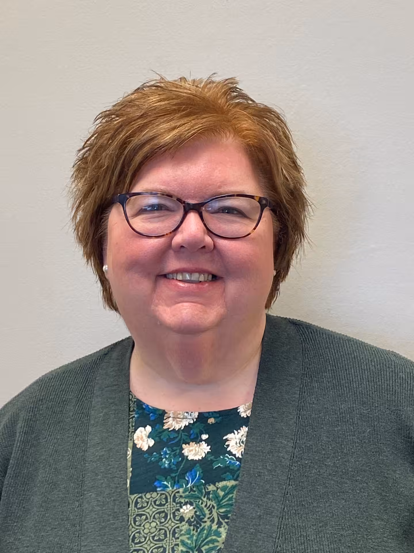 A woman with short, light brown hair and glasses smiles at the camera. She is wearing a dark green cardigan over a green floral-patterned top and is standing in front of a plain light-colored wall.