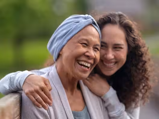 Two women sit close together on a bench, smiling and laughing. One woman wears a headscarf and the other has curly hair, with her arm lovingly around the other’s shoulder. The background is green and blurred.