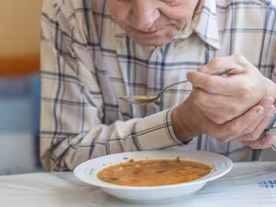 An elderly person in a plaid shirt eating soup from a white bowl with a spoon, seated at a table.
