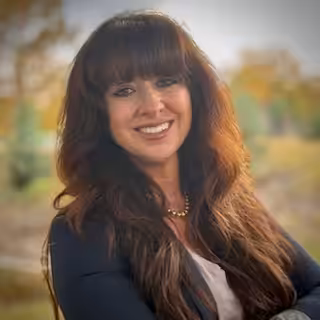 A woman with long brown hair and bangs smiles while standing outdoors. She is wearing a navy blazer, white top, and a gold necklace, with greenery and autumn trees blurred in the background.