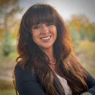 A woman with long brown hair and bangs smiles while standing outdoors. She is wearing a navy blazer, white top, and a gold necklace, with greenery and autumn trees blurred in the background.