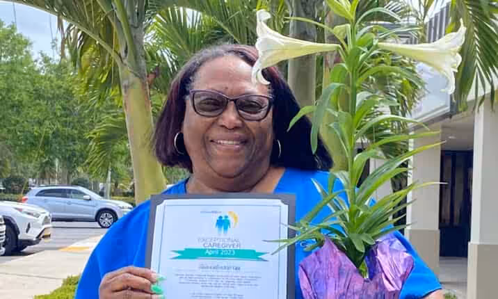 A smiling woman wearing glasses and a blue shirt holds a Caregiver of the Month April 2023 certificate and a potted plant with white flowers, standing outside near palm trees and parked cars.