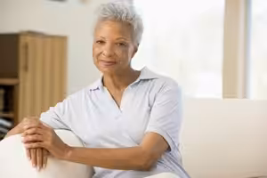 An older woman with short gray hair, wearing a light blue collared shirt, sits on a white sofa in a brightly lit room, smiling gently at the camera.