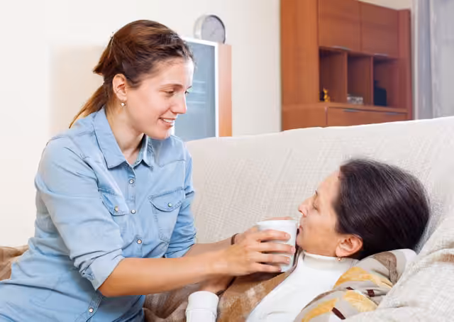 A young woman in a blue shirt helps an older woman lying on a couch by offering her a cup to drink from. The setting appears to be a cozy living room.