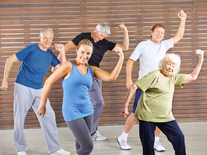 A group of five people of different ages, including seniors, are exercising together indoors, smiling and striking energetic, flexed-arm poses in workout clothes.