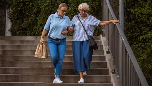 A younger woman helps an older woman walk down outdoor stairs, holding her arm for support. The younger woman carries paper bags, and both are dressed casually, surrounded by greenery.