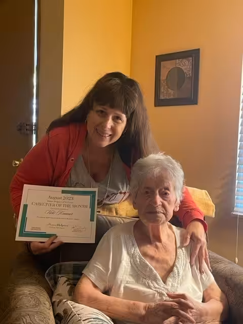 A woman with long brown hair smiles and holds a Caregiver of the Month certificate while standing beside an elderly woman seated in a chair, both in a warmly lit room with yellow walls and framed art.