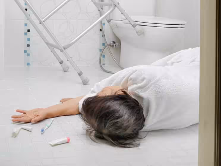 An older person in a white robe is lying on the bathroom floor near a toilet, with toiletries scattered nearby and a walker in the background, suggesting a possible fall.