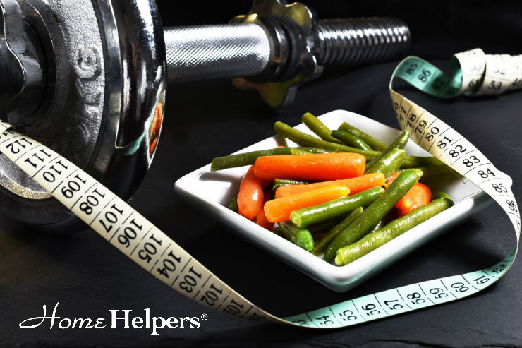 A dumbbell, a measuring tape, and a small plate of green beans and baby carrots are arranged on a dark surface. The Home Helpers logo appears in the corner, suggesting a focus on health and wellness.