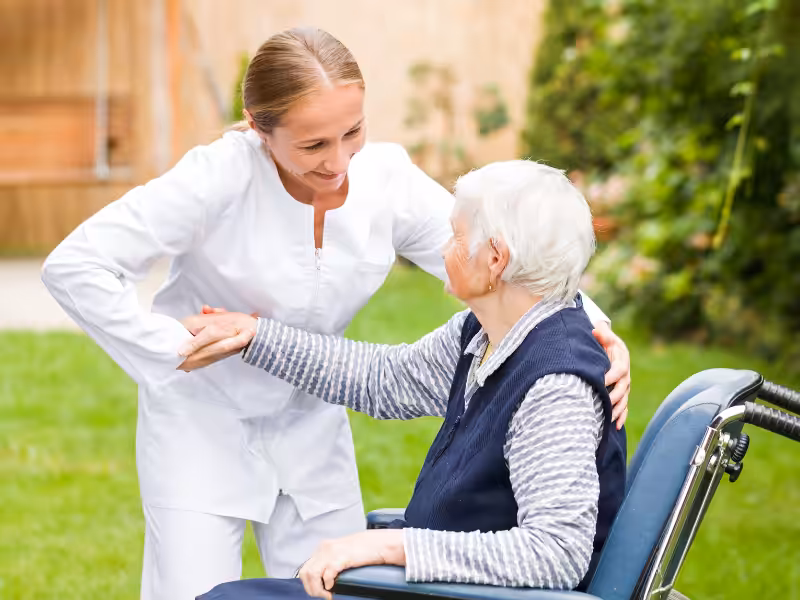 A caregiver in white helping an elderly woman in a wheelchair outdoors, smiling and holding her hand in a supportive manner. Green grass and plants are visible in the background.