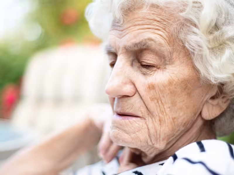Elderly woman with white hair and closed eyes, wearing a striped shirt, sitting outdoors with a contemplative expression.