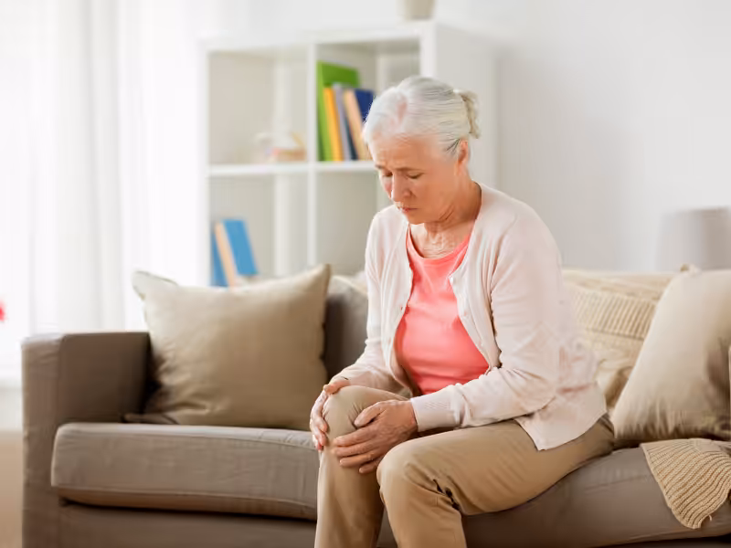 An elderly woman with gray hair sits on a beige sofa, holding her knee with a pained expression, suggesting discomfort or pain. A bookshelf and cushions are visible in the background.