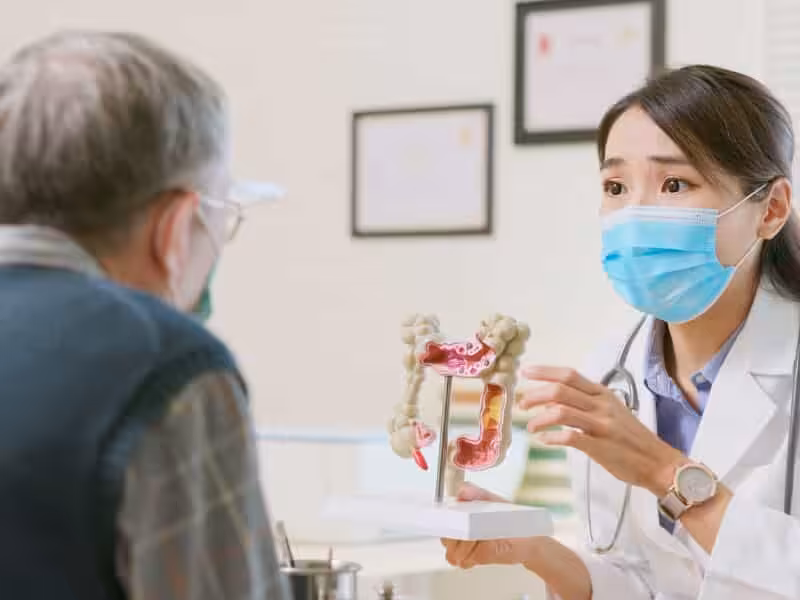A doctor wearing a mask explains a medical model of the human colon to an older patient during a consultation in a clinic.