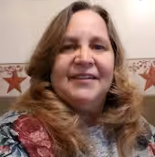 A woman with long, light brown hair and a floral top smiles at the camera. The background features a wall with a decorative border and red star ornaments.