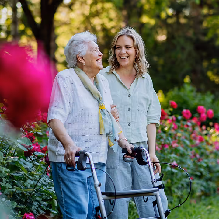 An elderly woman using a walker and a younger woman walk together in a garden with blooming pink flowers. Both are smiling and enjoying each other’s company on a sunny day.