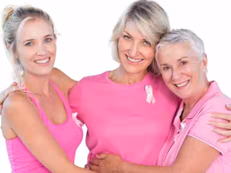 Three women wearing pink tops are standing close together, smiling. The woman in the middle has a pink ribbon pinned to her shirt, symbolizing breast cancer awareness. The background is white.