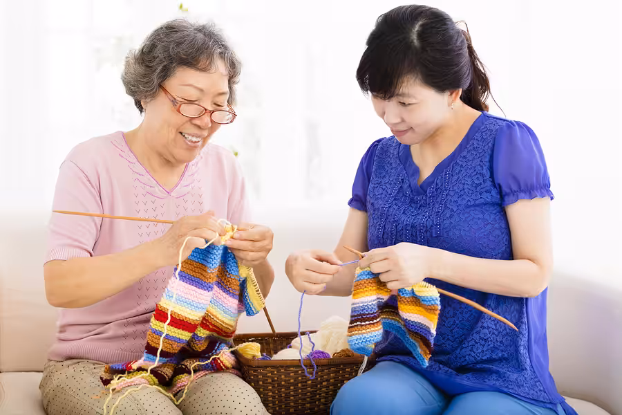 Two women sit on a couch, smiling as they knit colorful striped fabric pieces together. A basket with yarn sits between them. They appear to be enjoying their time crafting.