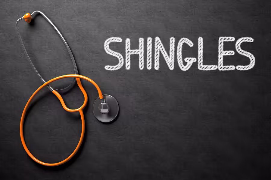A stethoscope with an orange tube lies on a blackboard surface next to the word SHINGLES written in large white chalk letters.