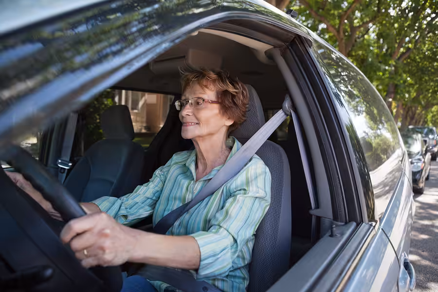 An older woman wearing glasses and a striped shirt smiles while driving a car on a sunny day. Trees and parked cars line the street outside her window.