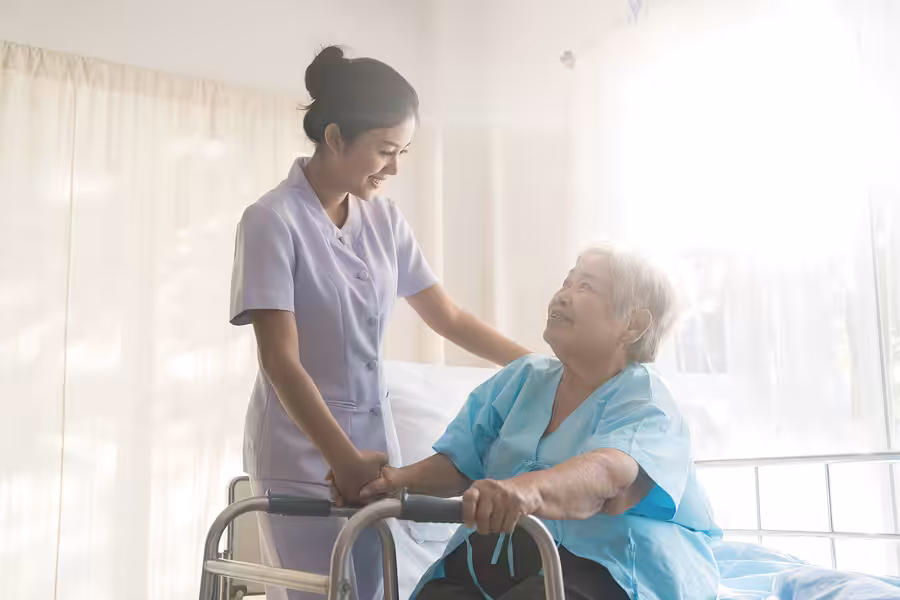 A nurse in a light purple uniform smiles and holds hands with an elderly woman using a walker, who is sitting on a hospital bed in a brightly lit room.