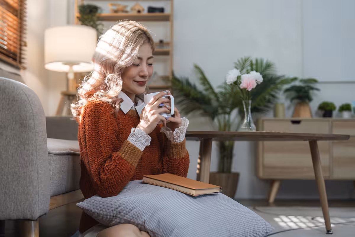 A woman with light hair sits on the floor by a sofa, holding a mug and smiling with her eyes closed. There is a pillow and a closed book in her lap. Sunlight filters in, and the room is cozy with plants and wooden furniture.