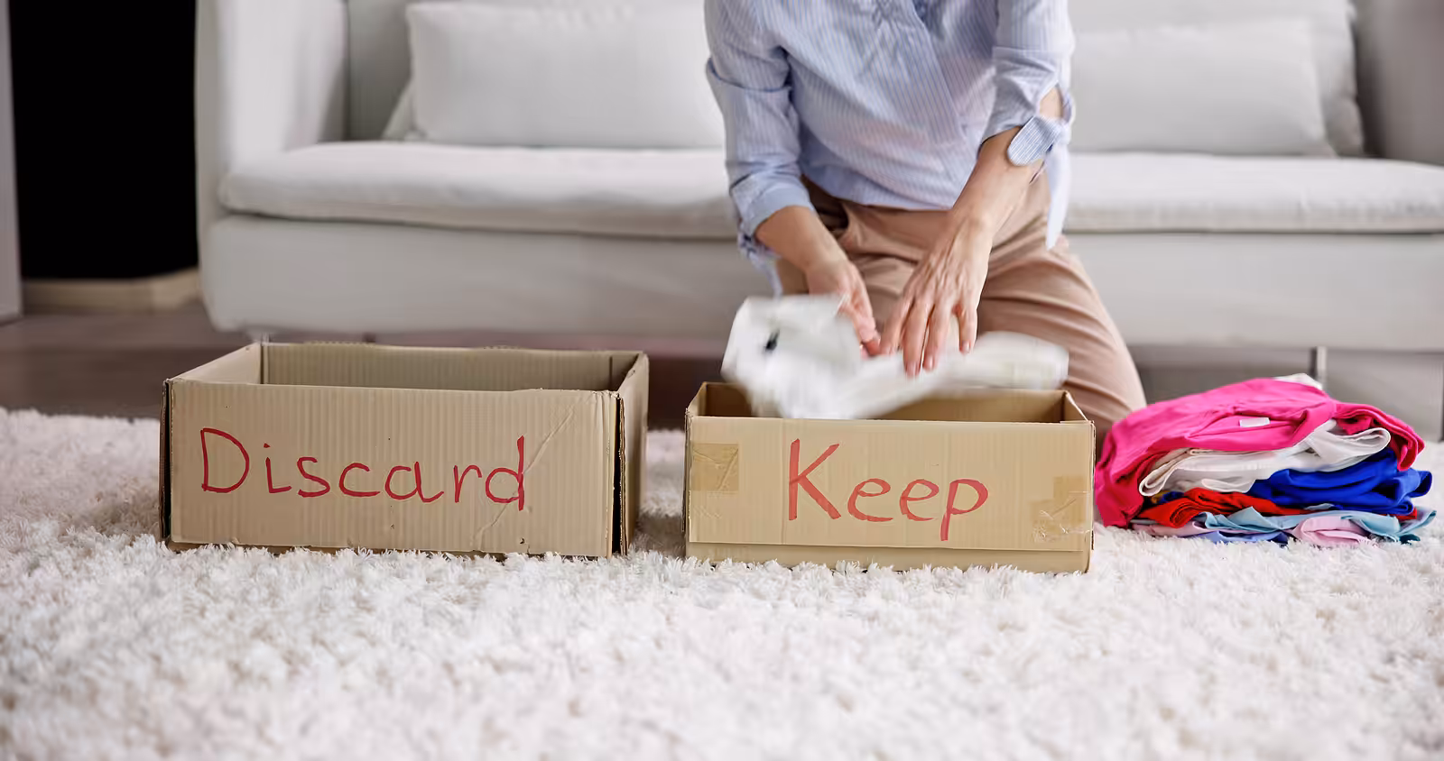A person sits on the floor in front of a sofa, sorting clothes into two cardboard boxes labeled Discard and Keep. Folded clothes are beside the boxes on a white carpet.
