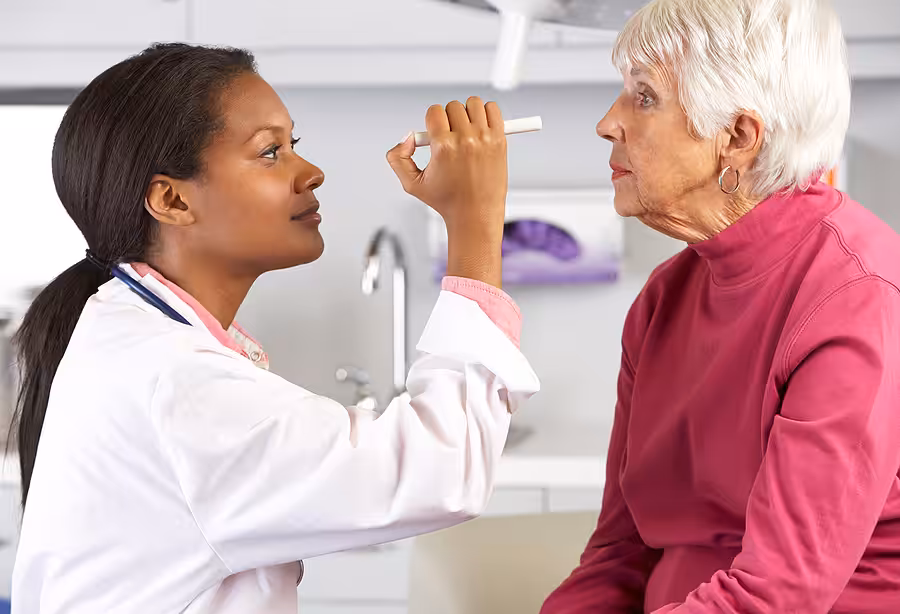 A healthcare professional examines an elderly womans eyes with a penlight in a medical office. The older woman sits on an exam table, looking ahead, while the doctor focuses on her eyes.
