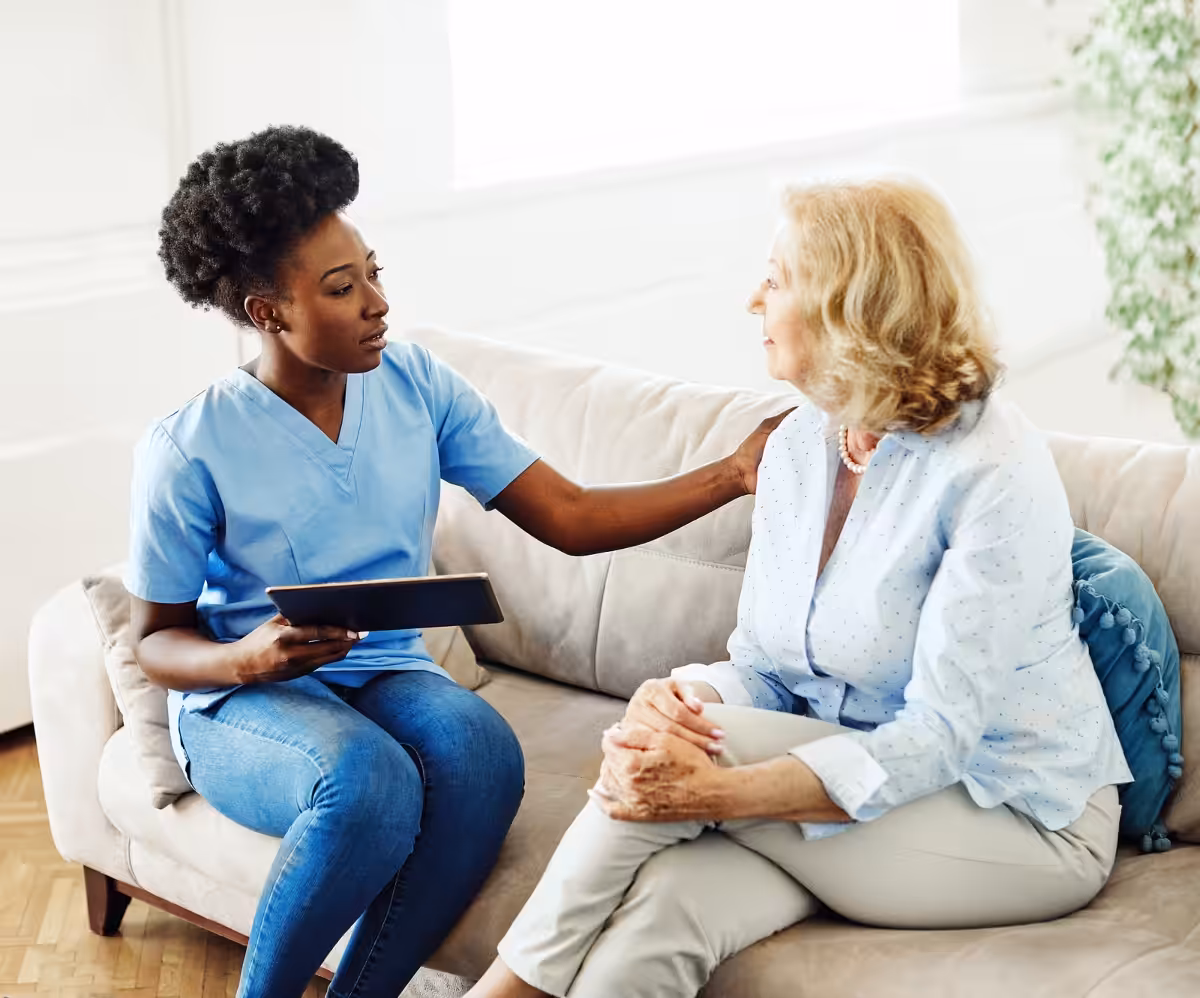 A nurse in light blue scrubs sits on a couch, holding a tablet and gently touching the shoulder of an older woman with light hair, offering comfort as they talk in a bright, home-like setting.