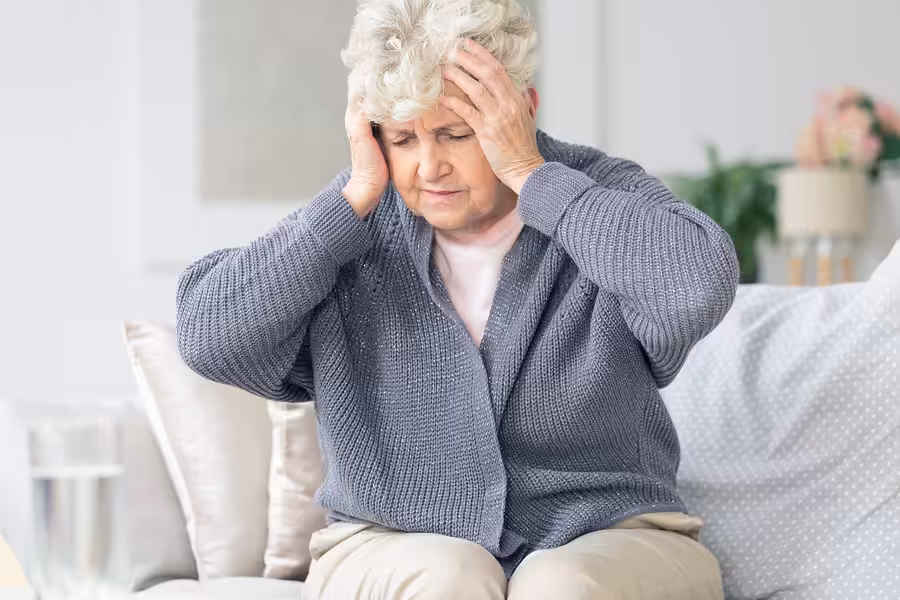An older woman sits on a sofa, holding her head with both hands and closing her eyes, appearing to be in pain or discomfort. She wears a gray sweater and light pants.