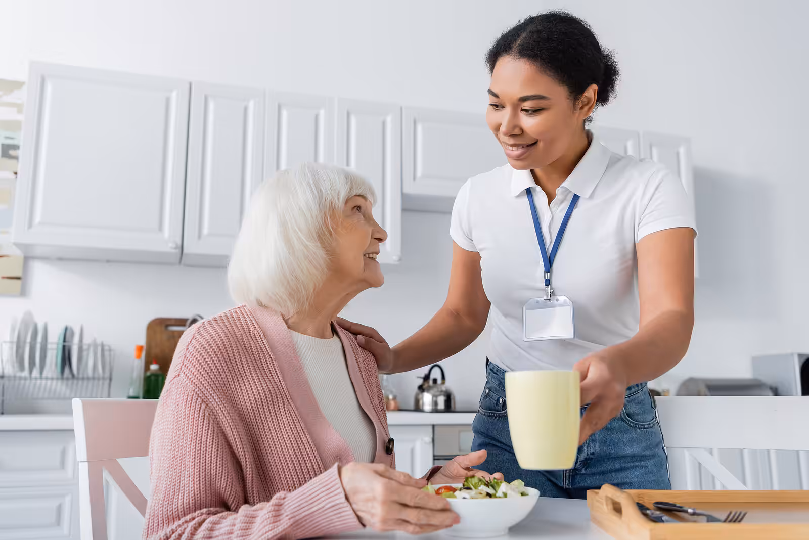 A young caregiver in a white shirt hands a yellow mug to an elderly woman in a pink cardigan, who is sitting at a kitchen table with a bowl of salad, smiling at each other.