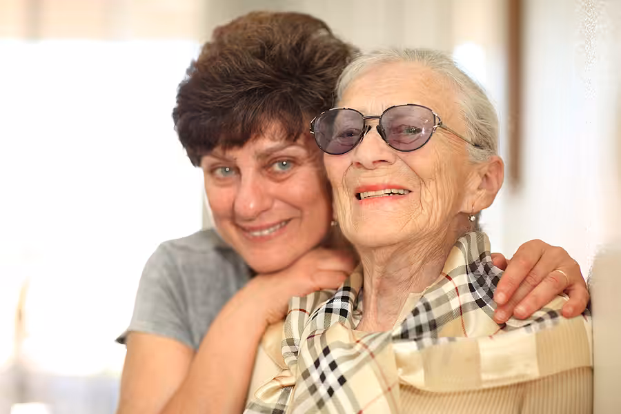 An older woman wearing sunglasses and a plaid scarf smiles while a younger woman stands close behind her, smiling with her arms resting on the older womans shoulders. Both appear happy and content.