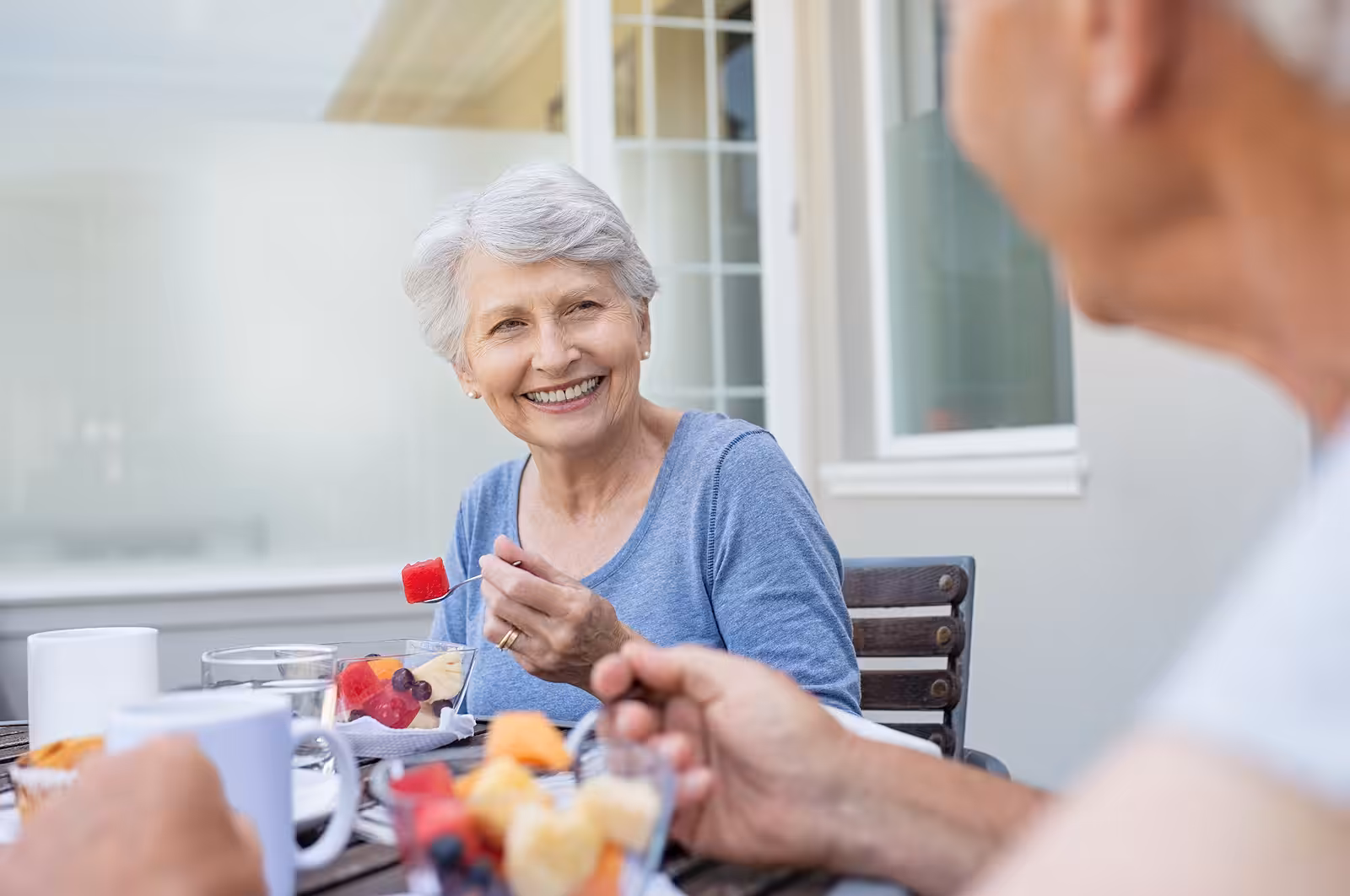 An older woman with short gray hair smiles while sitting at an outdoor table, holding a fork with fruit. Another person is blurred in the foreground. Plates of fruit and cups are on the table.