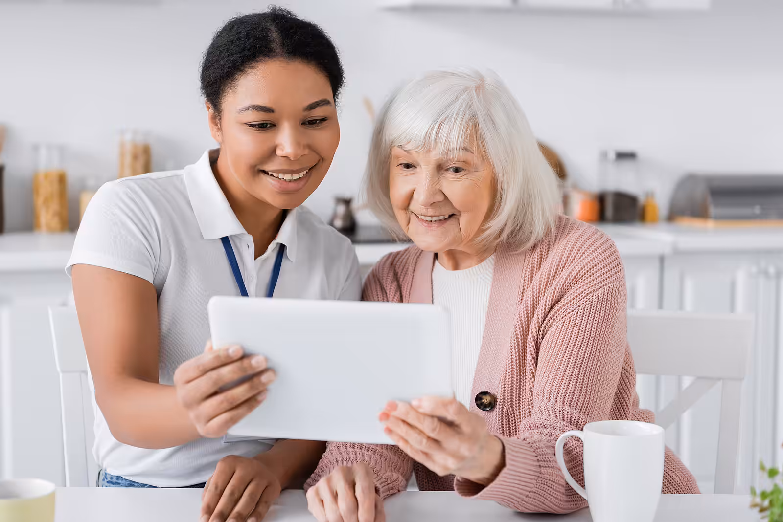 A young woman and an elderly woman sit together at a table, smiling while looking at a tablet. They appear to be enjoying each others company in a bright kitchen setting.