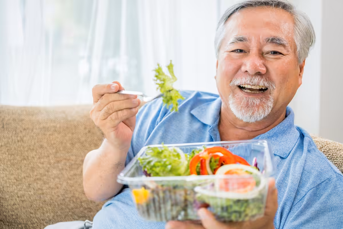 Smiling older man with gray hair and beard sits on a couch, holding a fork and a plastic container filled with fresh salad, including lettuce, tomatoes, and peppers. Bright natural light shines through a window behind him.