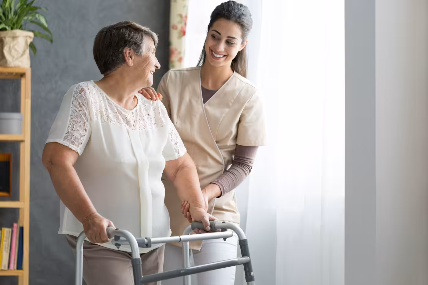 A smiling caregiver assists an elderly woman using a walker near a sunlit window, both appearing happy and engaged in a supportive, caring interaction.