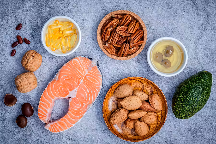 A variety of healthy fat sources displayed on a gray surface, including salmon, walnuts, pecans, almonds, avocado, olives in a bowl, and a small bowl of yellow fish oil capsules.