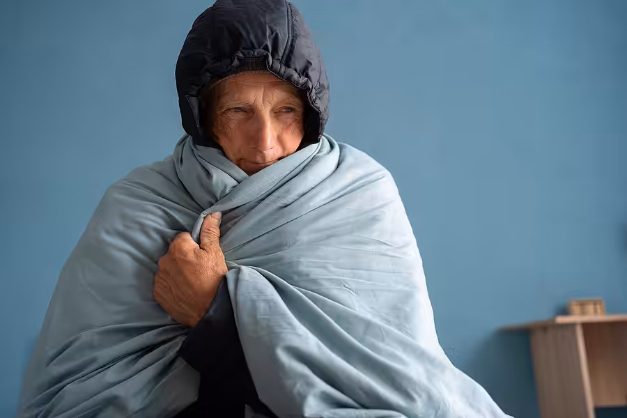 An older person wearing a dark hooded jacket sits wrapped in a light blue blanket, looking off to the side, with a plain blue wall and a small table in the background.