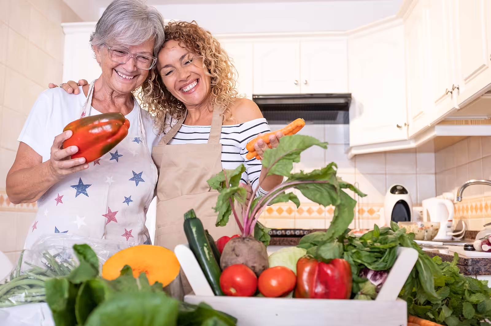 Two women in a kitchen smile while holding fresh vegetables, surrounded by a variety of colorful produce on the counter, including carrots, peppers, zucchini, and leafy greens.