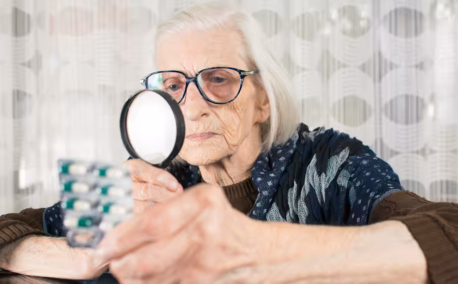 An elderly woman with glasses uses a magnifying glass to read the label on a blister pack of pills, sitting indoors near a window with sheer curtains in the background.