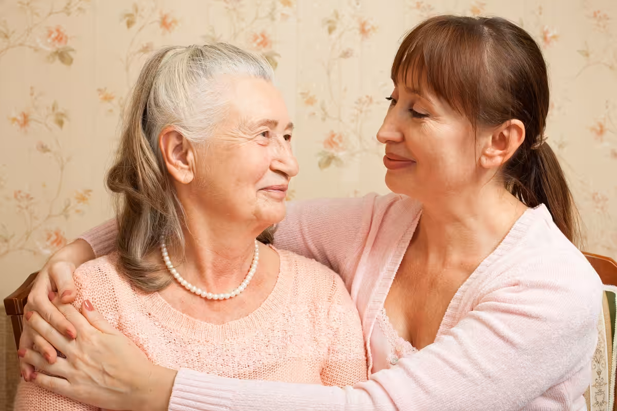 An older woman and a younger woman sit close together, smiling and embracing warmly. Both wear light pink sweaters in a cozy, vintage room with floral wallpaper.