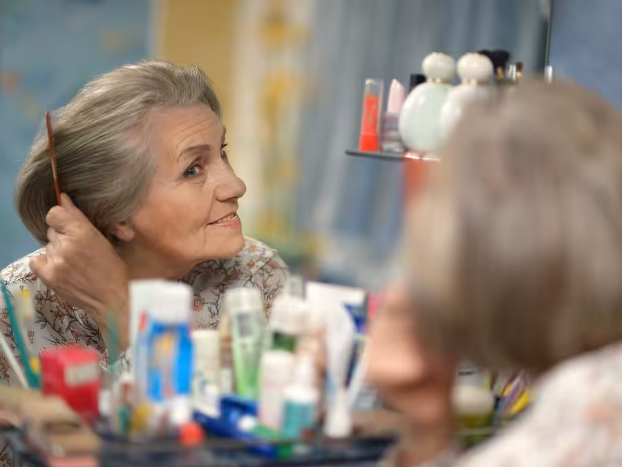 An elderly woman with gray hair is smiling and combing her hair while looking into a mirror. Various toiletries and cosmetic items are visible on the counter in front of her.
