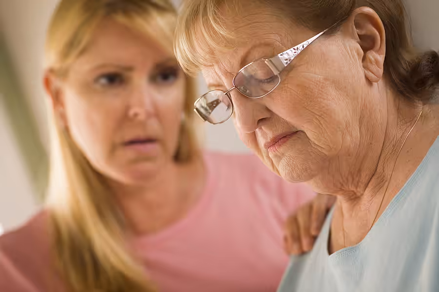 A younger woman gently comforts an older woman wearing glasses, who appears sad or deep in thought. The scene suggests care and support in a moment of emotional difficulty.