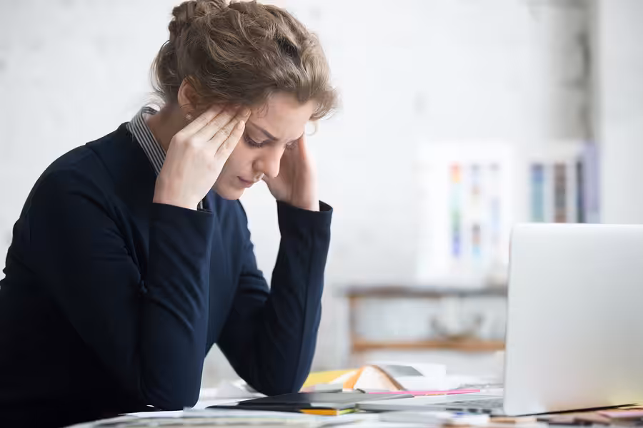 A woman sits at a desk with her hands on her temples, appearing stressed or tired, surrounded by papers, folders, and a laptop in a bright office setting.