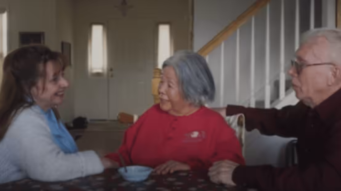 Three older adults sit together at a table in a cozy home, smiling and talking. A staircase and a front door are visible in the background, and a small dish sits on the patterned tablecloth.