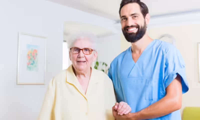 A smiling elderly woman wearing glasses stands next to a bearded male caregiver in blue scrubs, both looking at the camera and holding hands in a bright, home-like setting.