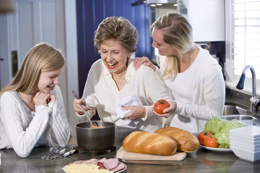 Three women of different ages smile and interact in a bright kitchen. One stirs a pot, while another holds tomatoes. Fresh bread, lettuce, and sandwich ingredients are on the counter. They look happy and engaged.