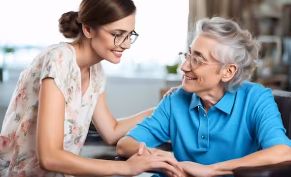 A young woman with glasses smiles and holds hands with an older woman in a wheelchair. Both women are looking at each other warmly indoors.