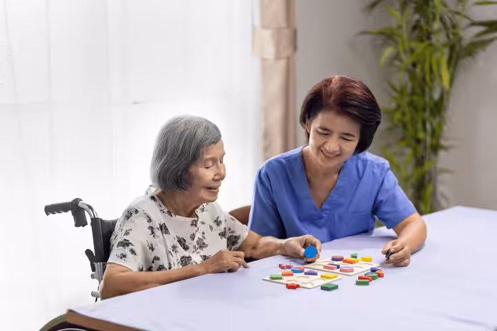 An elderly woman in a wheelchair and a caregiver in blue scrubs sit at a table, smiling and playing a colorful board game together in a bright room with a plant in the background.