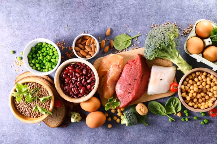 Assorted healthy foods including raw meat, fish, eggs, broccoli, peas, chickpeas, red beans, almonds, leafy greens, cherry tomatoes, and slices of whole grain bread arranged on a gray surface.