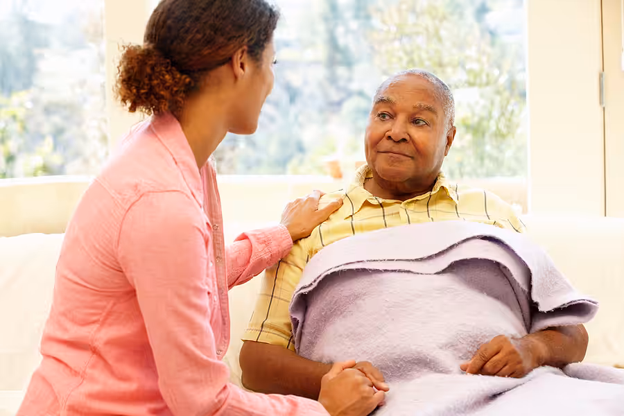 A young woman sits beside an older person wrapped in a blanket, gently holding their hand and touching their shoulder, offering comfort and support in a bright room.